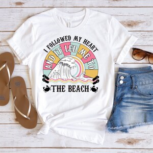May include: White t-shirt with a colorful graphic that reads "I followed my heart and it led me to the beach." The design includes a wave, sun, and beach elements. The image also shows denim shorts, sunglasses, and flip-flops.