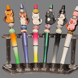 May include: A set of six decorative pens with cat-themed toppers. Each pen features a different cat design, colorful beads, and a gradient-colored barrel. The pens are displayed in a clear acrylic pen holder.