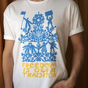 May include: A white short-sleeve t-shirt featuring a blue graphic print of stylised figures, floral motifs, and a central trident symbol. Below the graphic, the text FREEDOM IS OUR TRADITON is printed in yellow capital letters on the front of the shirt.