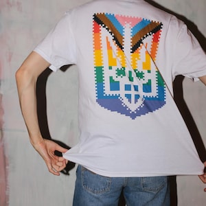 May include: White t-shirt featuring a pixelated design on the back. The design incorporates a shield shape with a rainbow color scheme and a cross symbol. The shirt is paired with blue jeans.