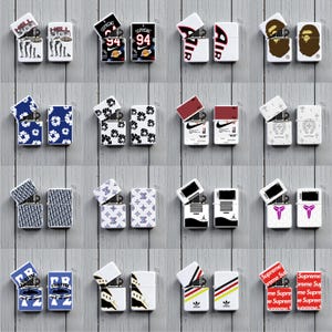 May include: A collection of sixteen metal flip-top lighters displayed in pairs, each featuring unique graphic designs including brand logos, floral patterns, and sports-themed prints. The lighters are shown both closed and with their lids open.