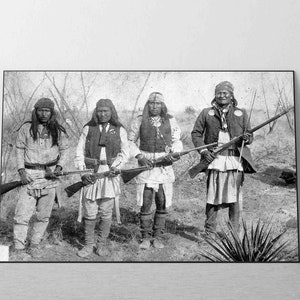 Op de afbeelding: Vier inheemse Amerikaanse mannen in traditionele kleding staan op een veld en houden geweren vast. Ze dragen gevederde hoofdtooien en hebben lang haar. De mannen staan voor een achtergrond van bomen en struiken.