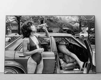 May include: A black and white photo of a woman in a swimsuit drinking from a bottle while sitting in a car. The car is parked on a street with houses in the background.