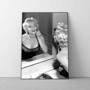 Op de afbeelding: Een zwart-witfoto van een vrouw die make-up aanbrengt voor een spiegel. Ze draagt een donkere jurk en heeft haar haar in een klassieke knot.