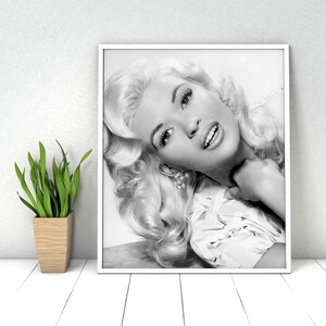 K&ouml;nnte beinhalten: Schwarzwei&szlig;-Portr&auml;tfotografie einer Frau mit blonden Haaren. Sie tr&auml;gt eine wei&szlig;e Bluse mit Blumenmuster und l&auml;chelt leicht.