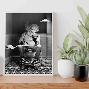 May include: A black and white photo of a man sitting on a toilet, smoking a pipe and writing on a roll of toilet paper. The photo is framed in a white frame and is hanging on a wall.