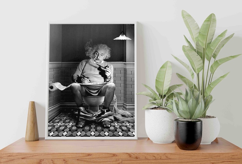May include: A black and white photo of a man sitting on a toilet, smoking a pipe and writing on a roll of toilet paper. The photo is framed in a white frame and is hanging on a wall.