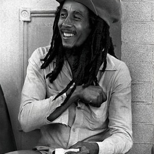 Puede incluir: Una foto en blanco y negro de un hombre con rastas largas que lleva una gorra y una camisa abotonada. Está sonriendo y sentado.