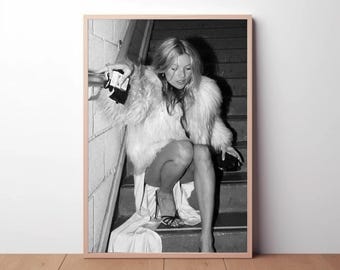 May include: A black and white photographic print featuring a person with long hair wearing a voluminous faux fur coat and a long skirt, sitting on a staircase. The person is holding a small dark handbag and wearing strappy high-heeled sandals.
