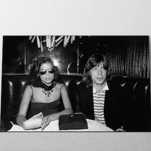 Puede incluir: Una fotografía en blanco y negro de una pareja sentada a una mesa en un restaurante. La mujer lleva un vestido negro y gafas de sol. El hombre lleva una camisa a rayas y una chaqueta.