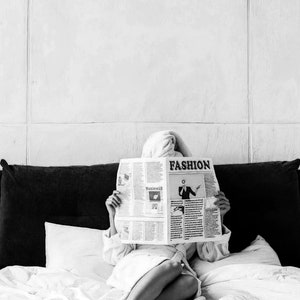 May include: A person in a white robe is sitting on a bed and reading a newspaper. The newspaper headline reads "FASHION".