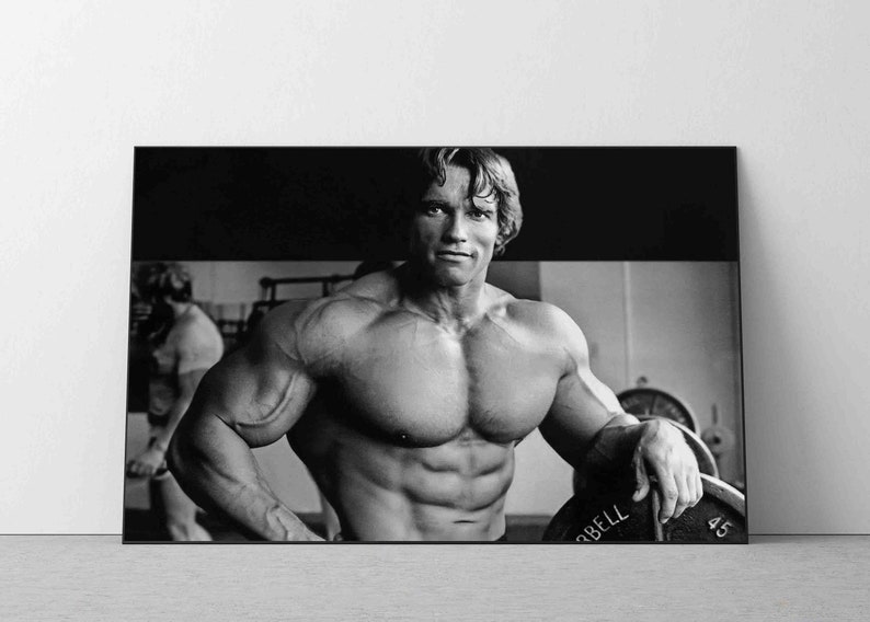 May include: Black and white photograph of a muscular man posing in a gym. He is wearing a pair of shorts and has a determined expression on his face. He is holding a weight plate in his right hand. The weight plate has the word "BELL" printed on it.