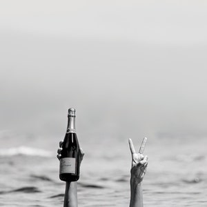 May include: A hand holding a bottle of champagne and another hand making a peace sign, both emerging from the water.