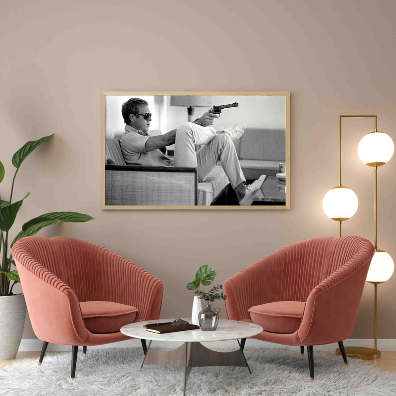 K&ouml;nnte beinhalten: Zwei rosa Sessel mit einem wei&szlig;en Marmor-Couchtisch vor einem Schwarzwei&szlig;foto eines Mannes, der auf einem Stuhl sitzt und eine Waffe h&auml;lt. Das Foto ist in Holz gerahmt.