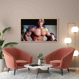 May include: A framed photograph of a muscular man posing in a gym. The man is shirtless and has a determined expression on his face. The photo is in a light wood frame and is hanging on a light pink wall.