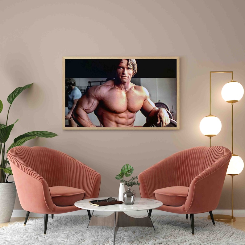 May include: A framed photograph of a muscular man posing in a gym. The man is shirtless and has a determined expression on his face. The photo is in a light wood frame and is hanging on a light pink wall.