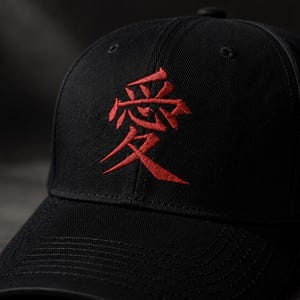 May include: A black baseball cap featuring a prominent red embroidered kanji character centered on the front panel. The cap is constructed from a structured fabric with a curved brim and visible stitching details along the visor and crown.