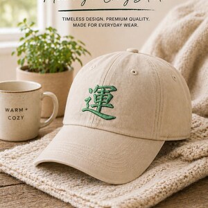 May include: A beige baseball cap with green embroidered Japanese characters. The cap is displayed on a textured beige surface, with a mug and small plant in the background. The text on the cap reads "Everyday Essential."