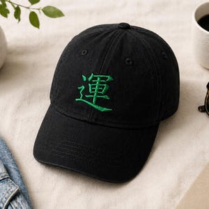 May include: Black baseball cap with green embroidered Japanese characters. The cap is on a light-colored surface, next to a white mug of coffee, sunglasses, and a pen.