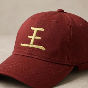 May include: A deep red baseball cap featuring a gold embroidered Chinese character on the front panel. The cap has a structured crown, a curved brim, and an adjustable metal buckle strap at the back for a secure fit, showcasing a classic design.