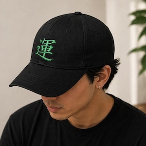 May include: Black baseball cap with a curved brim. The cap features green embroidered Japanese characters on the front. The cap is made of a soft material and has a structured fit.