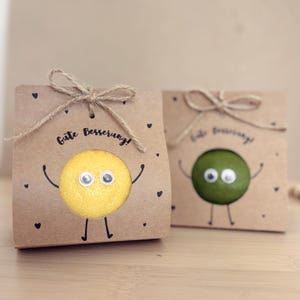 May include: Two small, brown paper gift bags tied with twine bows. One bag features a yellow lemon with googly eyes and arms, the other a green lime. Both have the phrase "Gute Besserung!" and small black heart details.