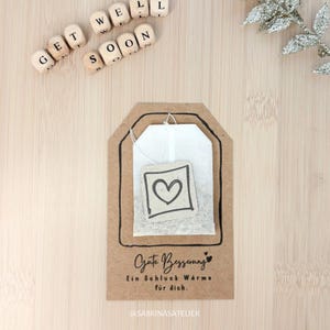 Op de afbeelding: Een kraft papieren theezakjestag met een witte theezakje erin, met een handgetekend hart. De tag heeft de woorden "Gute Besserung" en "Ein Schluck Wärme für dich" erop gedrukt. Houten letterblokken spellen "GET WELL SOON".