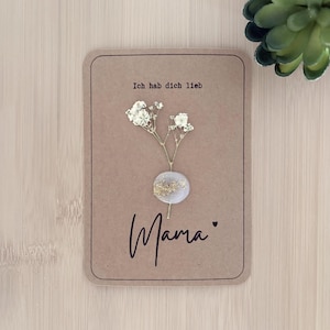 Peut inclure: Carte de vœux artisanale avec un fond marron. La carte présente le texte "Ich hab dich lieb" en haut et "Mama" en cursive en bas. Une petite branche de fleurs blanches est attachée à la carte.