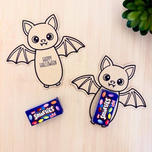 Puede incluir: Dos figuras de murciélago de cartón marrón claro troqueladas con contornos negros y ojos grandes. Un murciélago tiene el texto "HAPPY HALLOWEEN" en su cuerpo. Una pequeña caja azul de caramelos Smarties Mini está junto a uno y dentro del otro.