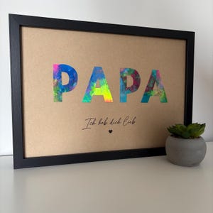 May include: Framed artwork with the word "PAPA" in colorful, abstract lettering. Below, the text "Ich hab dich lieb" is written in cursive, with a small heart symbol. The frame is black, and the background is tan.