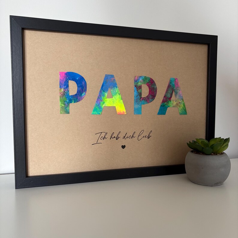 May include: Framed artwork with the word "PAPA" in colorful, abstract lettering. Below, the text "Ich hab dich lieb" is written in cursive, with a small heart symbol. The frame is black, and the background is tan.