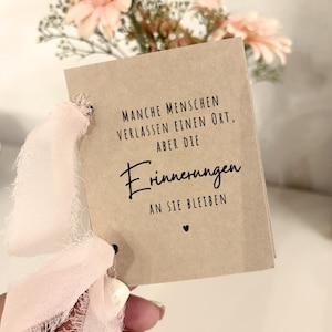Könnte beinhalten: Ein rechteckiger, hellbrauner Kartonanhänger mit einem ausgefransten, hellrosa Band, das durch ein Loch auf der linken Seite gezogen ist. Der Text lautet: MANCHE MENSCHEN VERLASSEN EINEN ORT, ABER DIE Erinnerungen AN SIE BLEIBEN, gefolgt von einem kleinen Herz.