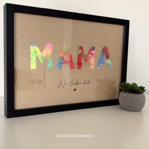 Puede incluir: Una obra de arte enmarcada con la palabra "MAMA" en letras coloridas y texturizadas. Debajo, el texto "Wir lieben dich" est&aacute; escrito en cursiva, con un peque&ntilde;o s&iacute;mbolo de coraz&oacute;n. El marco es negro y el fondo marr&oacute;n claro.
