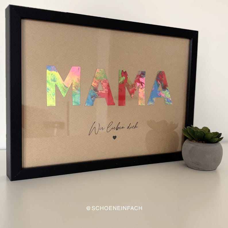 Puede incluir: Una obra de arte enmarcada con la palabra "MAMA" en letras coloridas y texturizadas. Debajo, el texto "Wir lieben dich" est&aacute; escrito en cursiva, con un peque&ntilde;o s&iacute;mbolo de coraz&oacute;n. El marco es negro y el fondo marr&oacute;n claro.