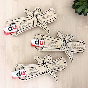 May include: Three light tan paper cutouts shaped like rolled diplomas tied with bows. Each holds a small chocolate bar with a white and red wrapper. The text reads HAST'S GESCHAFFT!, ABI 2026, HERZLICHEN GLÜCKWUNSCH, and ENDLICH BACHELOR! in black ink.