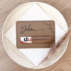 May include: A brown card with the words "Schön, dass du da bist" in black script, a small chocolate bar, and a sprig of dried grass, all arranged on a white napkin and a cream-colored plate.