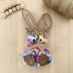 Puede incluir: Una bolsa de celofán transparente llena de caramelos coloridos, con forma de conejito. El conejito tiene ojos saltones, una corona floral y huellas de patas. La bolsa está atada con hilo de bramante, y las palabras "Frohe Ostern" están escritas con bloques de letras de madera.