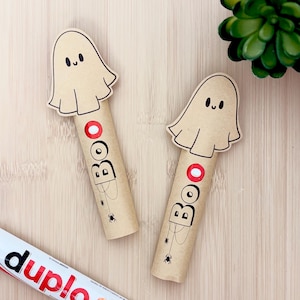 Op de afbeelding: Twee cilindrische kartonnen kokers versierd met een beige spookuitsparing aan de bovenkant. Elke koker bevat het woord BOO in zwarte letters met een rode cirkel in plaats van de letter O, vergezeld van kleine zwarte spinnenillustraties en een Duplo-reep.