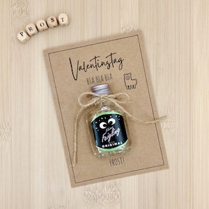 Puede incluir: Una pequeña botella de licor con una etiqueta negra y verde, atada con hilo, se encuentra sobre una tarjeta marrón. La tarjeta dice "Valentinstag" y "BLA BLA BLA" con una ilustración de llama. Bloques de letras de madera deletrean "PROST".