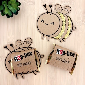 Può includere: Tre sagome di carta a forma di ape in cartoncino marrone. Una presenta strisce glitterate dorate e un volto sorridente. Due fungono da involucri per oggetti rettangolari, stampati con il testo "hap-bee BIRTHDAY" e "TO YOU".