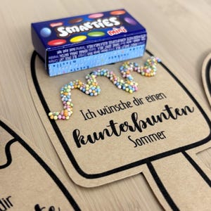 Puede incluir: Un recorte de papel marr&oacute;n con forma de helado y borde negro, que presenta el texto "Ich w&uuml;nsche dir einen kunterbunten Sommer" y una l&iacute;nea ondulada de peque&ntilde;as perlas de az&uacute;car multicolores. Una caja azul de Smarties Mini descansa encima.