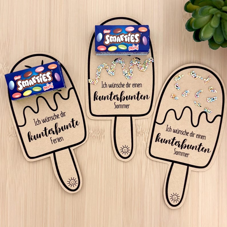 Puede incluir: Tres tarjetas de cart&oacute;n marr&oacute;n claro con forma de helado, contornos negros y un peque&ntilde;o icono de sol en el palo. Dos tarjetas tienen una caja azul de Smarties mini, una tiene virutas de colores. Texto: "Ich w&uuml;nsche dir kunterbunte Ferien" o "Sommer."