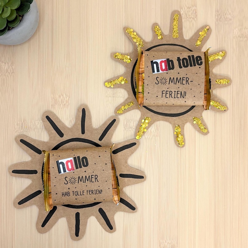 May include: Two sun-shaped cardboard cutouts with black hand-drawn rays and gold glitter accents. Each cutout features a wrapped chocolate bar in the center with printed text. One reads "hallo SOMMER HAB TOLLE FERIEN!" and the other "hab tolle SOMMER-FERIEN!"