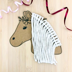 Op de afbeelding: Een paardenkop van knutselpapier met een bloemenkrans en een witte macramé manen. De paardenkop is versierd met een zwart oog en een bruine stip voor de neus. Roze en bordeauxrode linten zijn op de achtergrond.