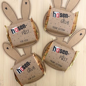 Peut inclure: Quatre barres de chocolat enveloppées dans du papier brun avec les mots "hasen-POST" et "FÜR DICH" imprimés dessus. Les barres sont placées sur des découpes en forme de lapin marron avec des détails de cœur noirs. Les mots "FROHE OSTERN!" sont également imprimés.