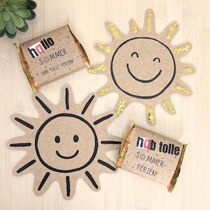 Könnte beinhalten: Zwei rechteckige Schokoladentafeln in braunem Papier mit dem Text "hallo SOMMER HAB TOLLE FERIEN!" und "hab tolle SOMMER-FERIEN!" sowie zwei sonnenförmige Pappaufsteller, einer mit schwarzen Linien und einer mit goldenen Glitzerakzenten.