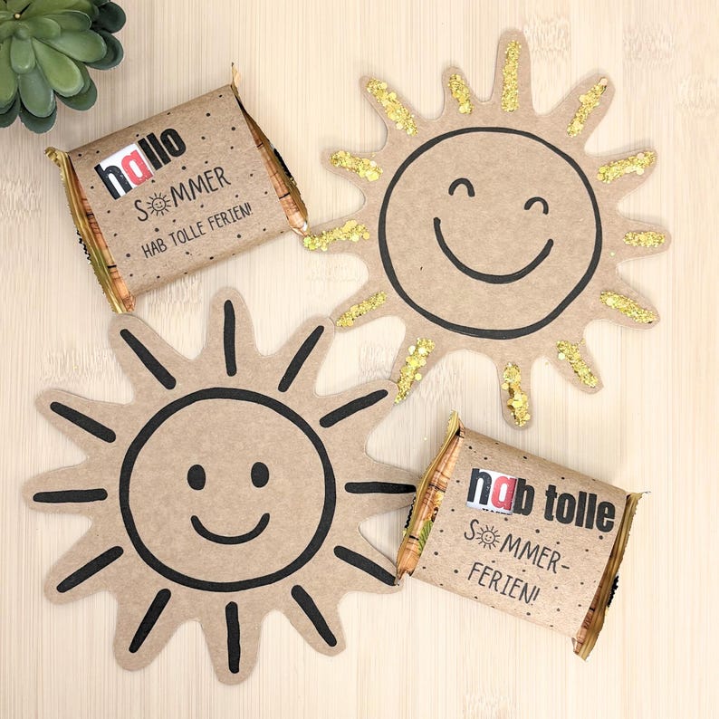 May include: Two rectangular chocolate bars wrapped in brown paper with the text "hallo SOMMER HAB TOLLE FERIEN!" and "hab tolle SOMMER-FERIEN!" alongside two sun-shaped cardboard cutouts, one with black ink lines and one with gold glitter accents.