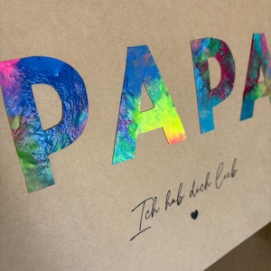 May include: A tan card with the word "PAPA" in large, colorful, abstract letters. Below, the phrase "Ich hab dich lieb" is written in cursive, with a small heart symbol.