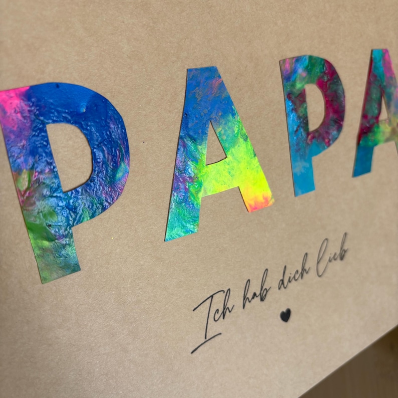 May include: A tan card with the word "PAPA" in large, colorful, abstract letters. Below, the phrase "Ich hab dich lieb" is written in cursive, with a small heart symbol.