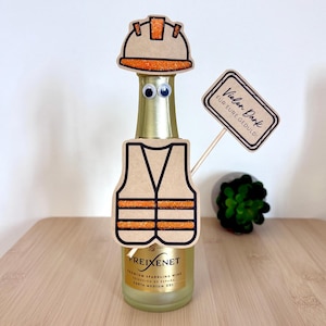 May include: A Freixenet sparkling wine bottle decorated with a paper construction worker outfit, including a hard hat and safety vest with orange glitter accents, googly eyes, and a sign on a stick that reads Vielen Dank FÜR EURE GEDULD!